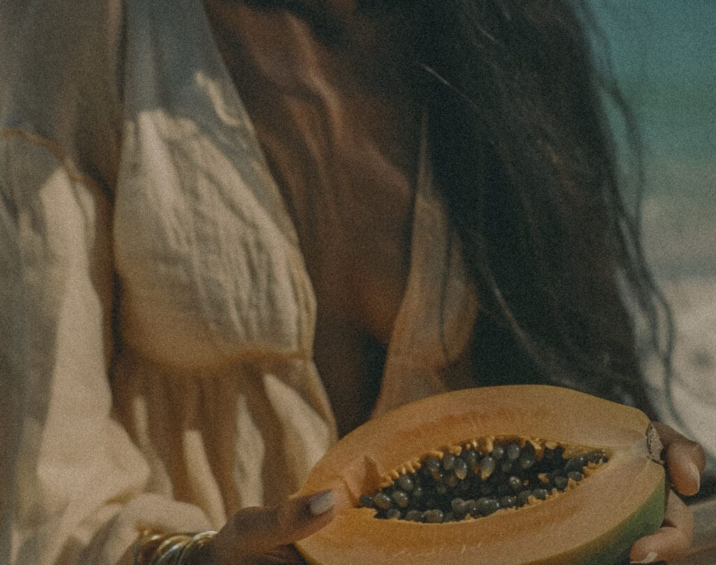 Close up shot of papaya in hands, woman with long dark hair wearing beige boho dress and golden bracelets, at the beach, captured in the style of Alasdair_Evans, natural lighting, Kodak film photo, pastel colors.