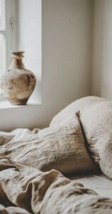 A close-up photo of linen bed sheets in a minimalist bedroom with window light and natural lighting. In the background there is an old vase on top of one pillow. Neutral colors provide calming vibes with an aesthetic background in the style of minimalism.